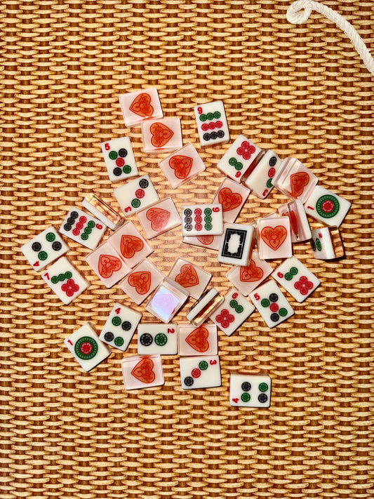 Warm white acrylic mahjong tiles scattered face up on rattan, showing dots, bams, cracks and dragon tiles alongside tile backs printed with the red heart logo.