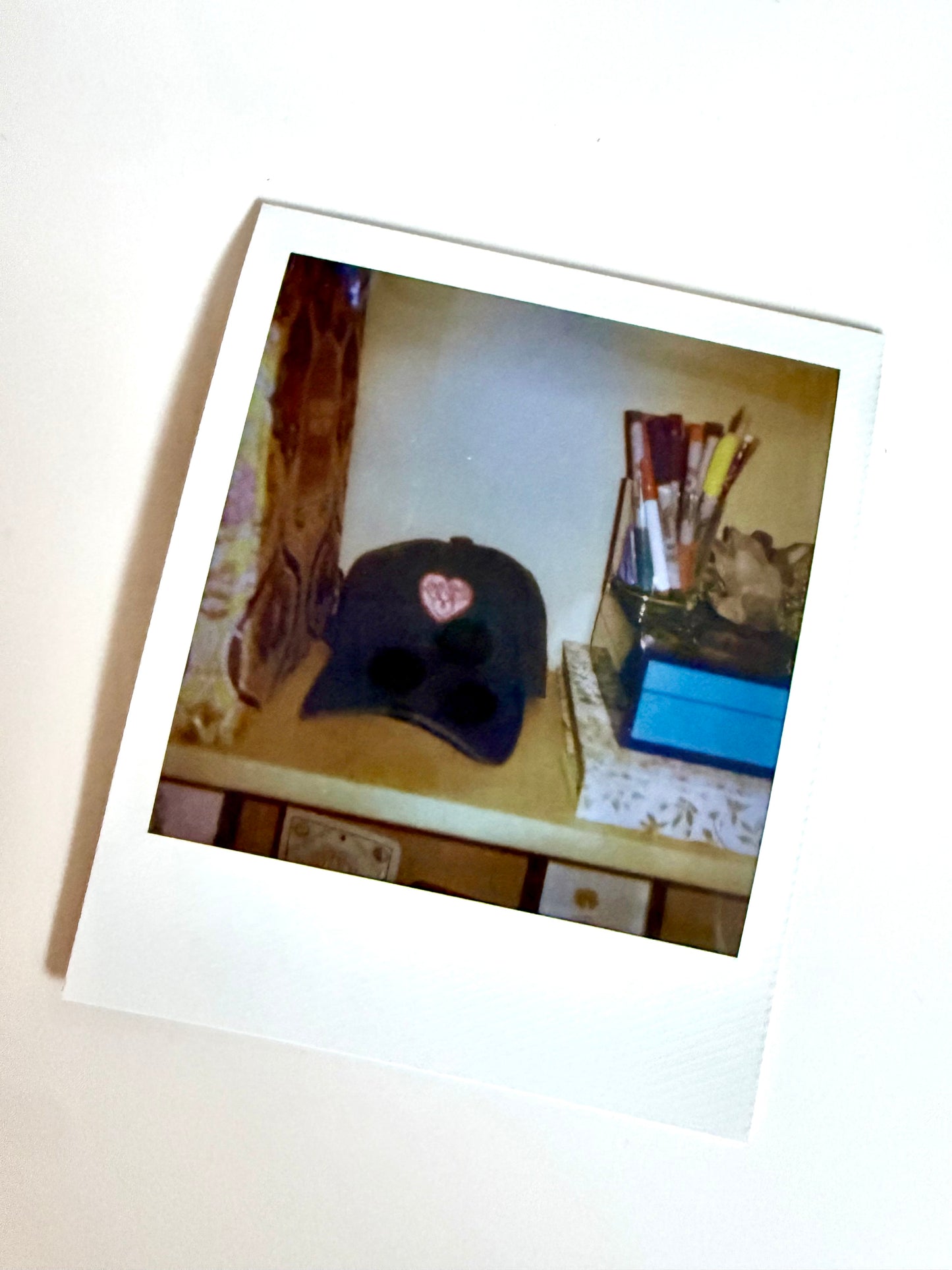 Polaroid photo of an embroidered heart-crest cap on a desk beside a jar of paintbrushes