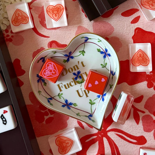 Two red logo dice with pink heart pips in a heart-shaped porcelain dish, surrounded by mahjong tiles on a pink floral mat