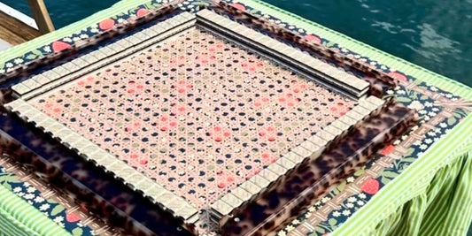A strawberry-print caned mahjong mat set with tortoiseshell racks and tiles, on a green striped skirted table at the water's edge