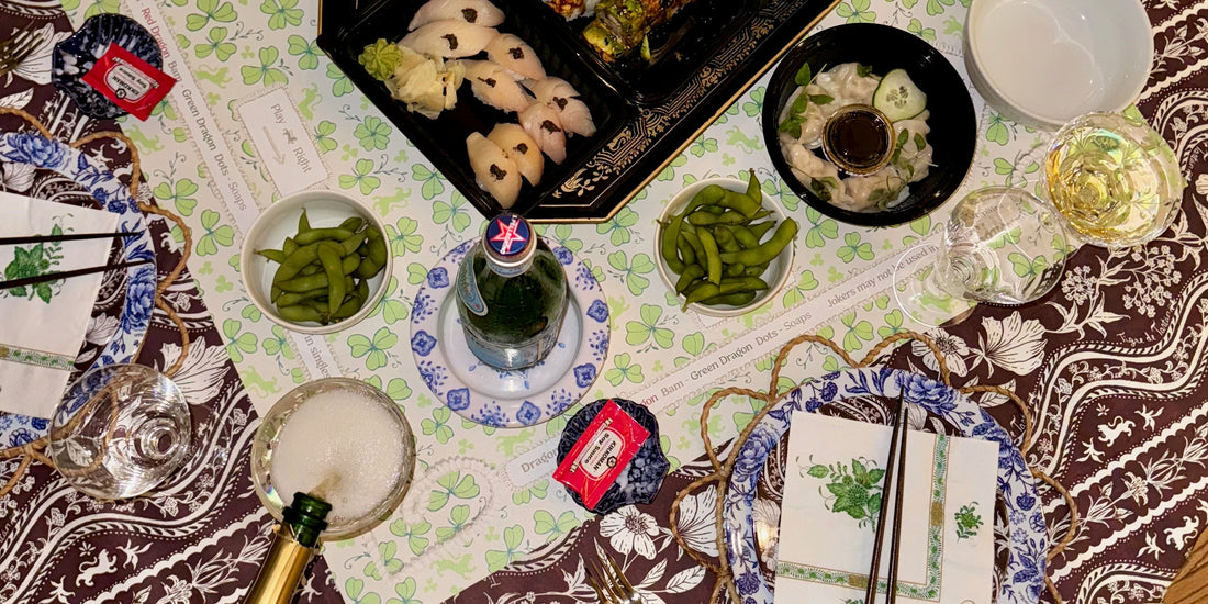 Overhead view of a mahjong table mid-evening: sushi, edamame, wine glasses and a champagne pour, on a printed Tigre de Tartán cloth