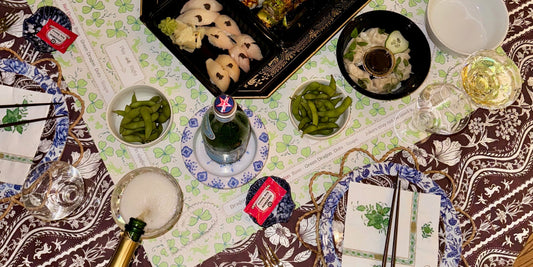 Overhead view of a mahjong table mid-evening: sushi, edamame, wine glasses and a champagne pour, on a printed Tigre de Tartán cloth