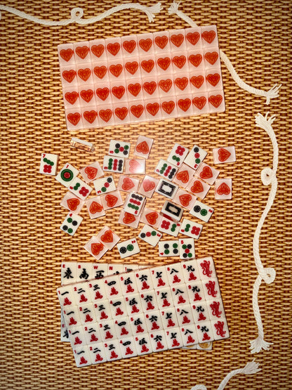 House Hearts warm white American mahjong tiles on a woven rattan surface, with a rack of tile backs showing the red Tigre de Tartán heart-and-tigers logo above racks of traditional character tiles.