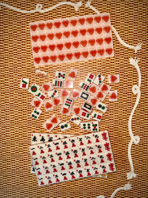 House Hearts warm white American mahjong tiles on a woven rattan surface, with a rack of tile backs showing the red Tigre de Tartán heart-and-tigers logo above racks of traditional character tiles.