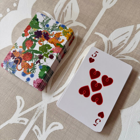 Jardín de Los Jefes playing cards: floral tiger-print deck beside the five of hearts with faux-stitched heart pips, on a linen tablecloth