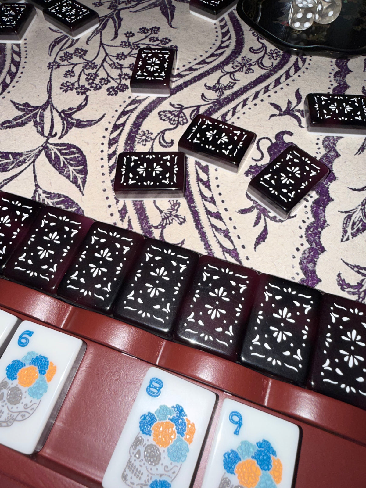 Oxblood mahjong rack holding black and white floral-pattern tiles, with loose tiles scattered on a purple floral tablecloth