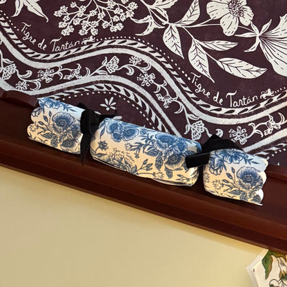Three Chinoiserie Bleu party crackers lined up on a dark wood mantel in front of a Domino Oxblood patterned backdrop with Tigre de Tartán script