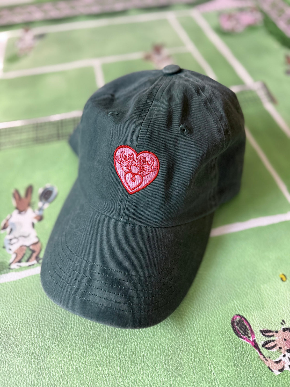 Hunter green baseball cap with pink embroidered House Hearts crest, on a green tennis-print fabric
