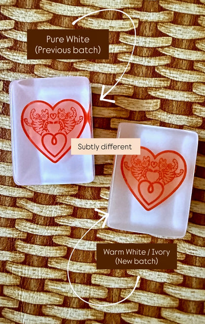Side-by-side comparison of two House Hearts tile backs: the previous pure white batch beside the new warm white and ivory batch, labeled subtly different.