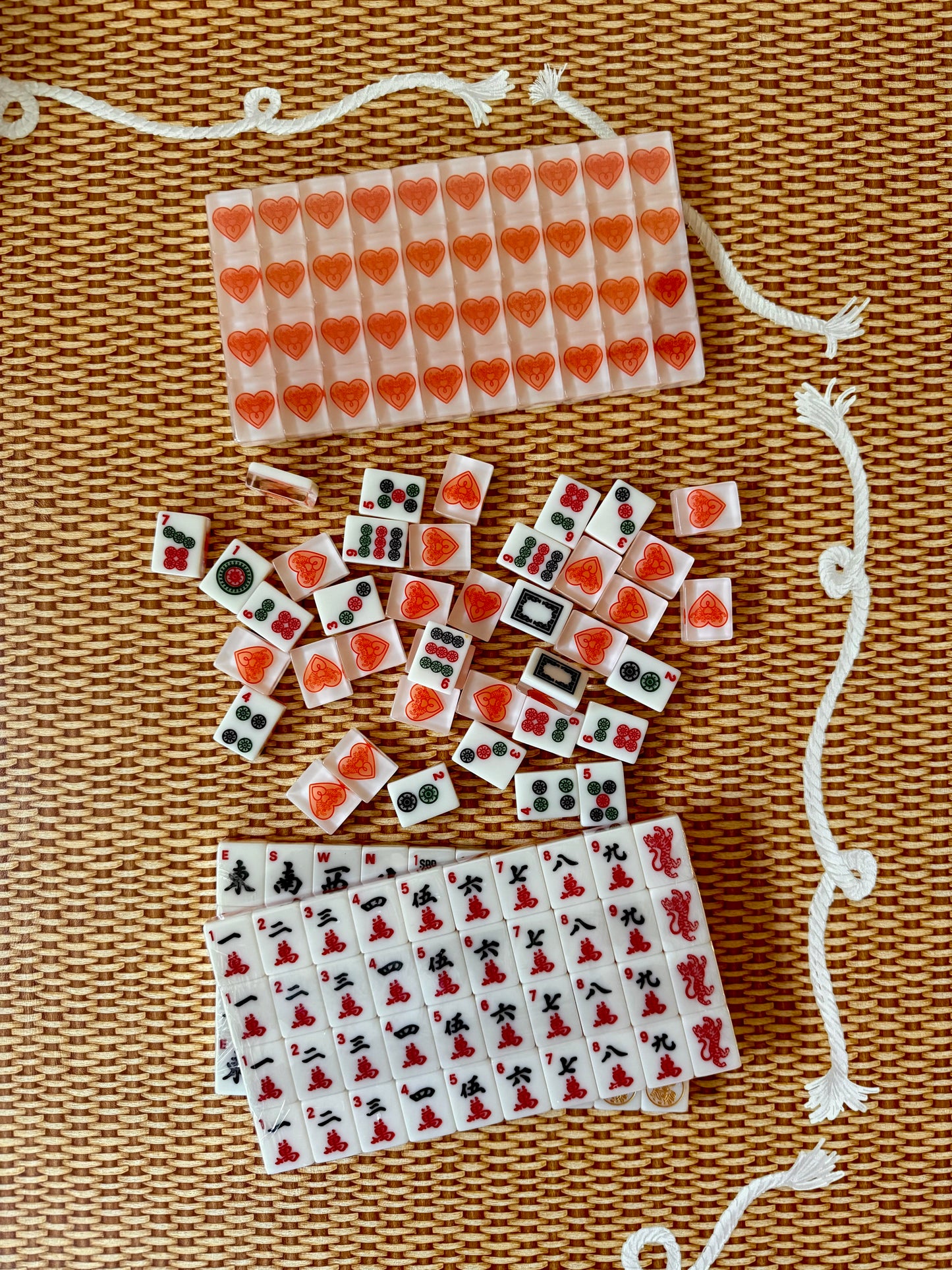 Warm white House Hearts mahjong tiles scattered above two racks of traditional Chinese character tiles in red and black, on woven rattan with a white rope cord.