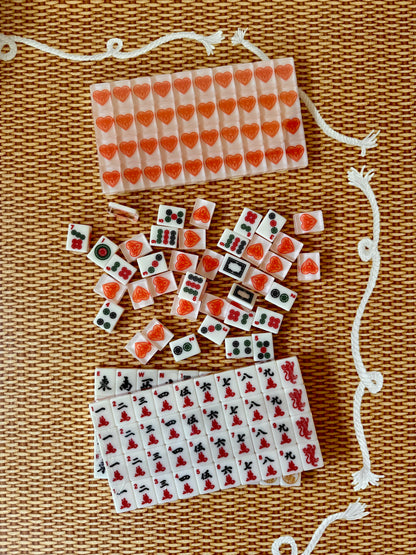 Warm white House Hearts mahjong tiles scattered above two racks of traditional Chinese character tiles in red and black, on woven rattan with a white rope cord.