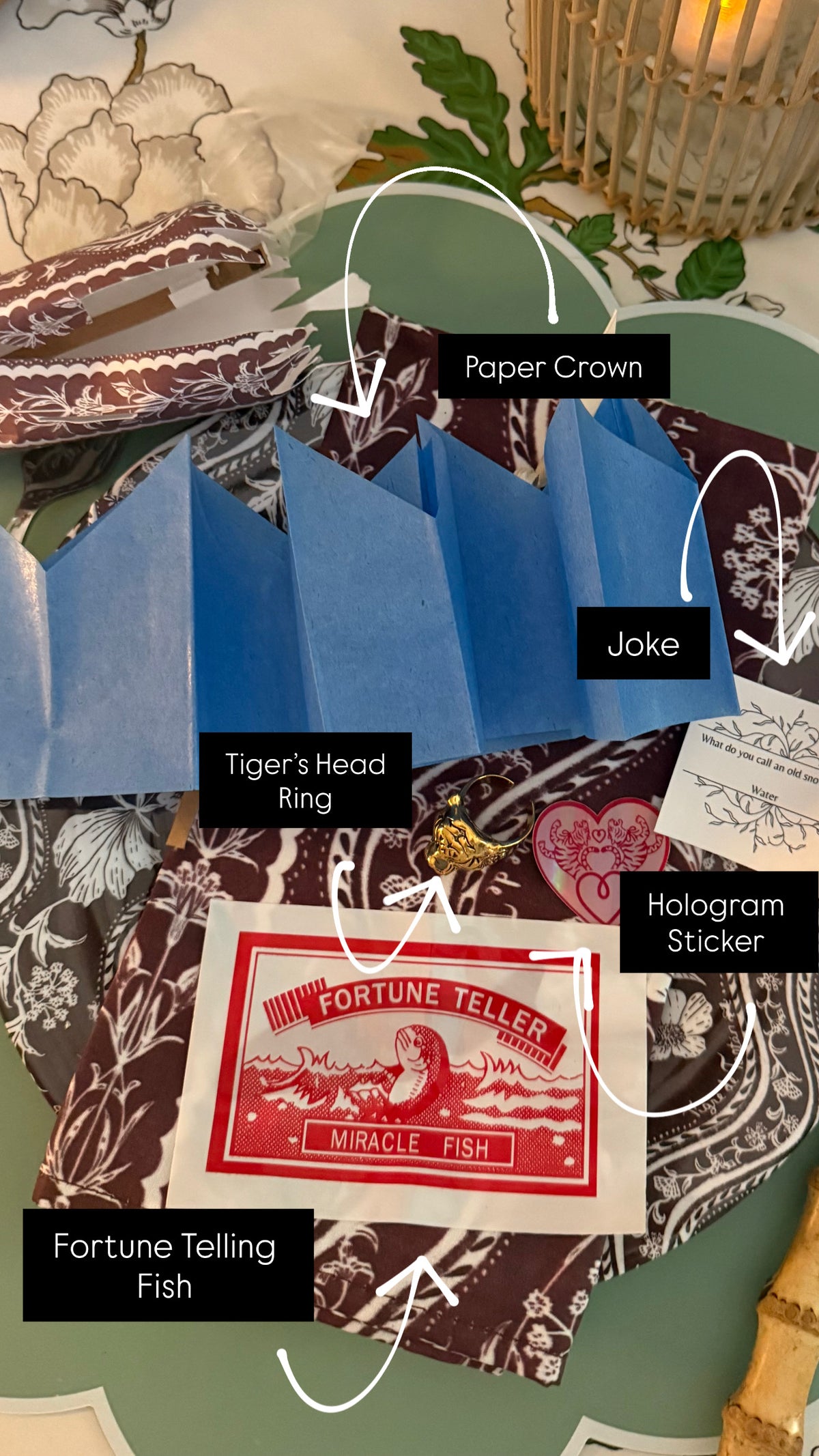 Annotated flat-lay of party cracker contents: paper crown, joke, tiger's head ring, hologram sticker and fortune-telling fish beside a Domino Oxblood cracker