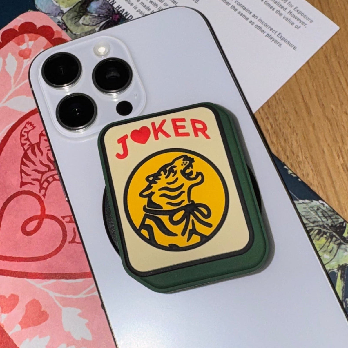 Joker phone grip with a yellow tiger Joker tile design and red JOKER lettering on the back of a silver iPhone