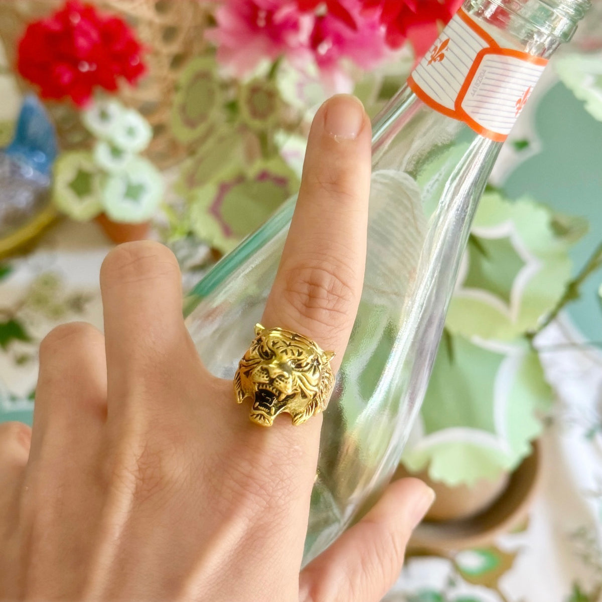 Hand wearing the gold tiger-head ring from the party cracker, holding a glass soda bottle over a floral tablecloth