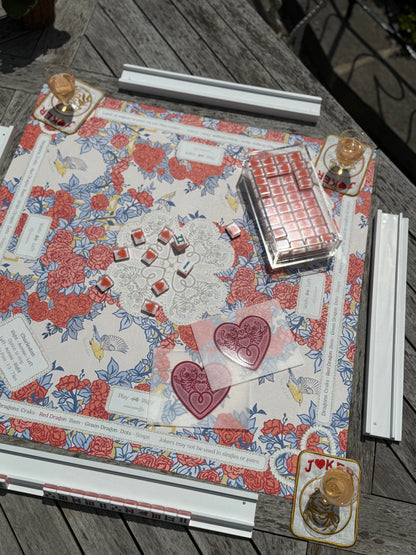 Four white racks around a red and blue floral mahjong mat with heart tiles, a pink tile set and drinks coasters