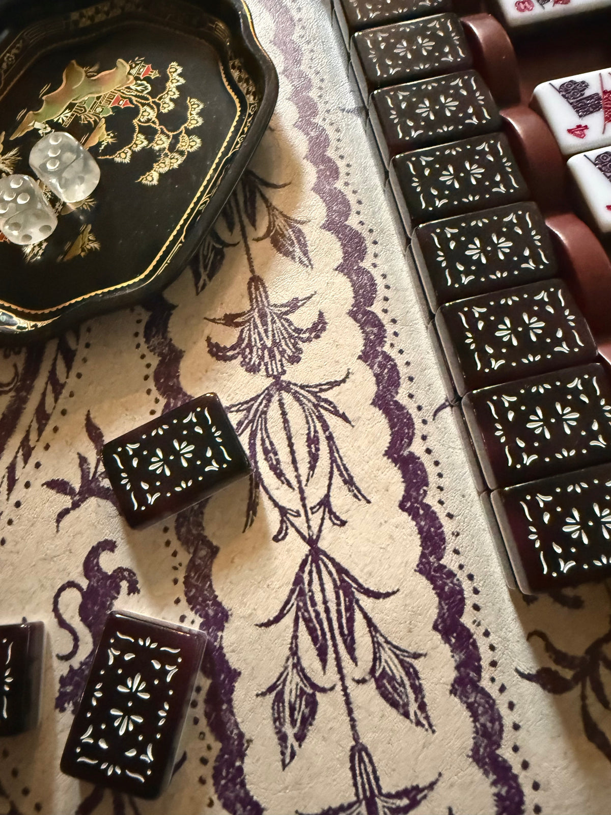 Oxblood mahjong rack edge with dark floral-pattern tiles, next to a lacquered dice tray with dice