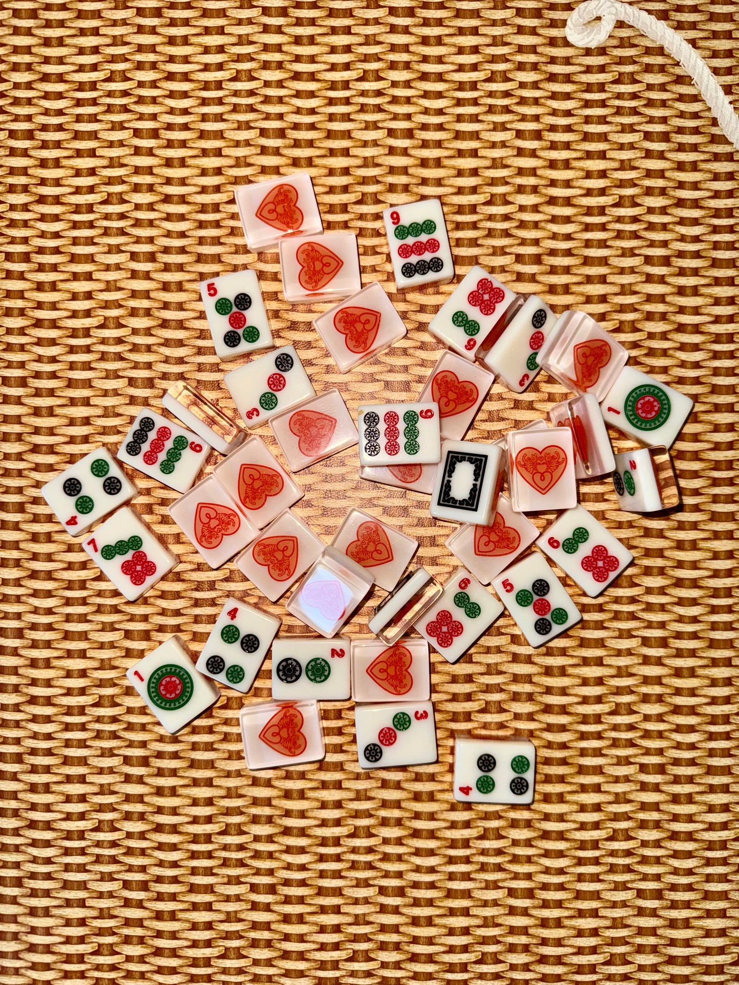 Warm white acrylic mahjong tiles scattered face up on rattan, showing dots, bams, cracks and dragon tiles alongside tile backs printed with the red heart logo.