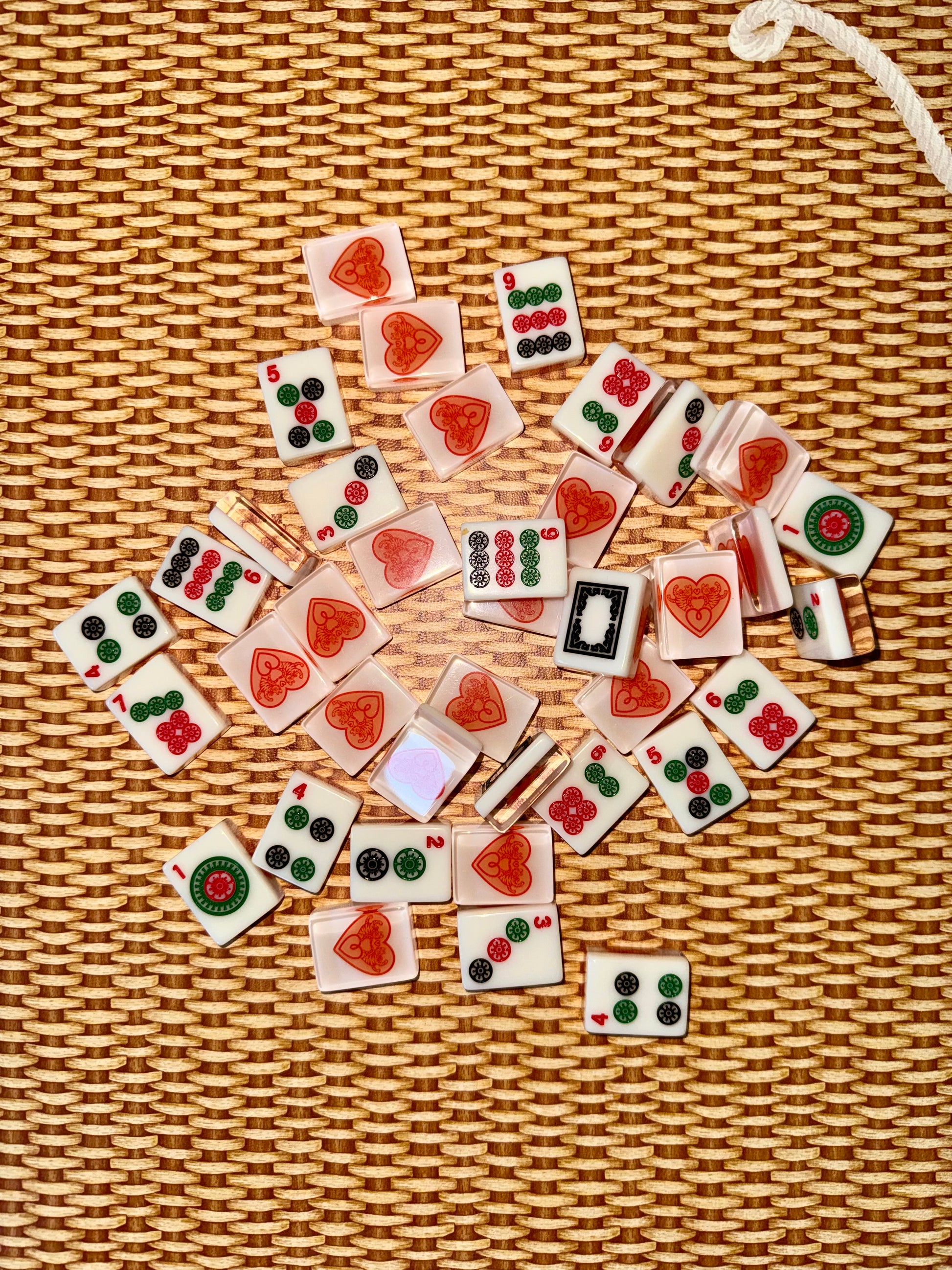 Warm white acrylic mahjong tiles scattered face up on rattan, showing dots, bams, cracks and dragon tiles alongside tile backs printed with the red heart logo.