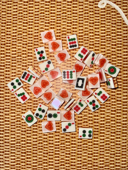 Warm white acrylic mahjong tiles scattered face up on rattan, showing dots, bams, cracks and dragon tiles alongside tile backs printed with the red heart logo.