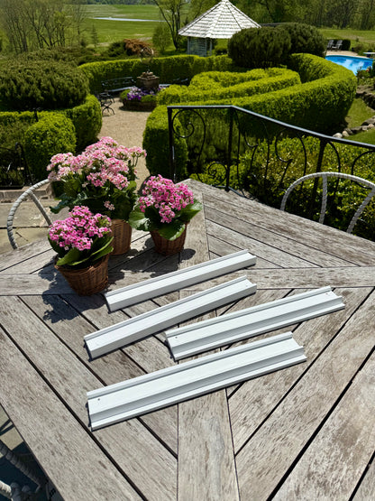 Four white beech wood mahjong racks on a weathered teak table beside potted pink flowers, garden in the background
