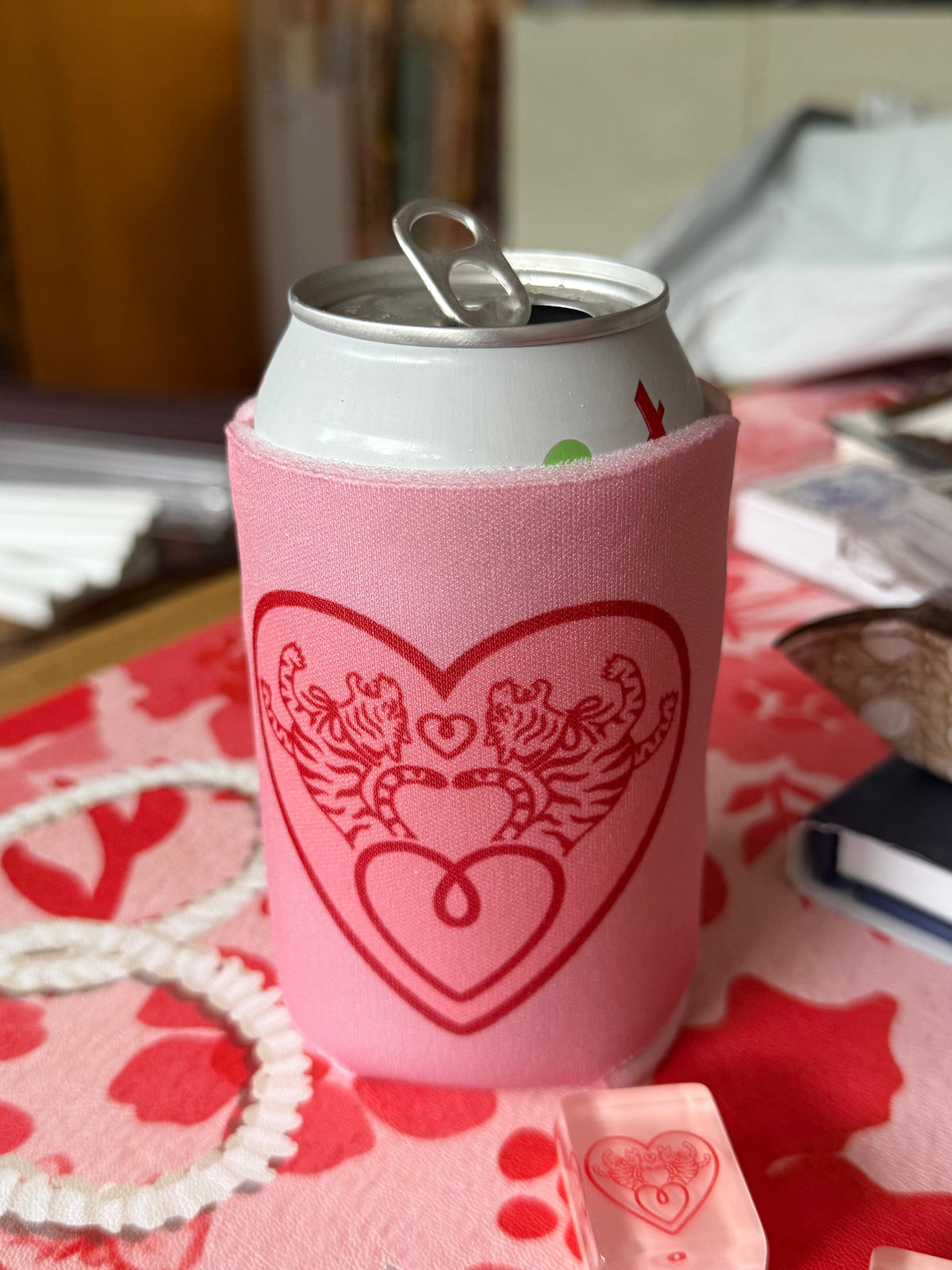 Pink foam can koozie with a geranium-red Tiger Heart print on a 12 oz can, sitting on a pink and red floral mahjong mat with a pearl necklace and a pink tile
