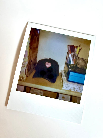 Polaroid photo of an embroidered heart-crest cap on a desk beside a jar of paintbrushes