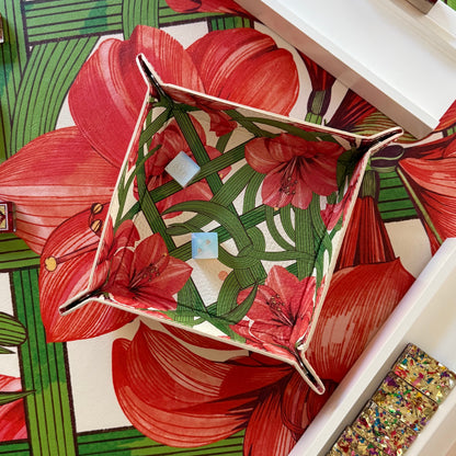 A styled flat-lay of a square fabric basket with a red floral and green leaf pattern, placed on top of a matching printed fabric runner. Several small patterned boxes and a tray of colorful glitter are arranged around it on a white surface.