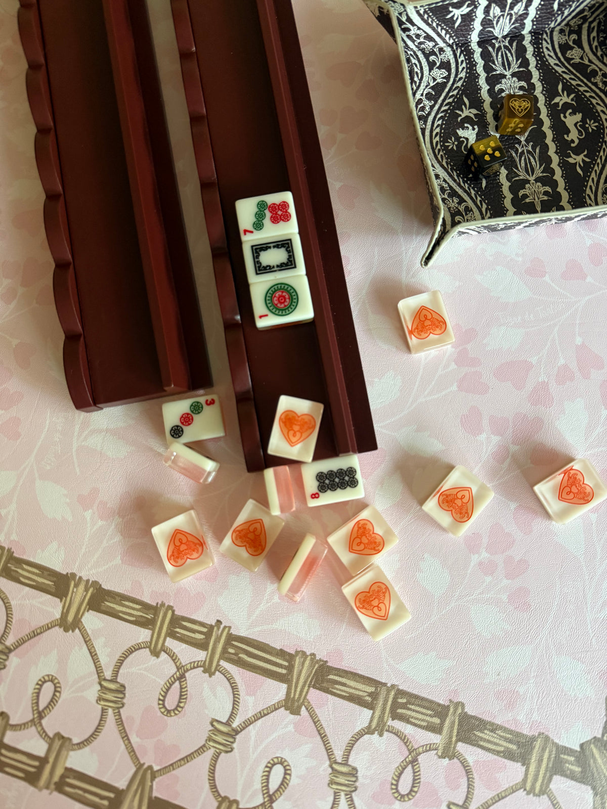 Two empty oxblood mahjong racks with cream heart-pattern tiles scattered nearby, on pink floral fabric with a dice tray