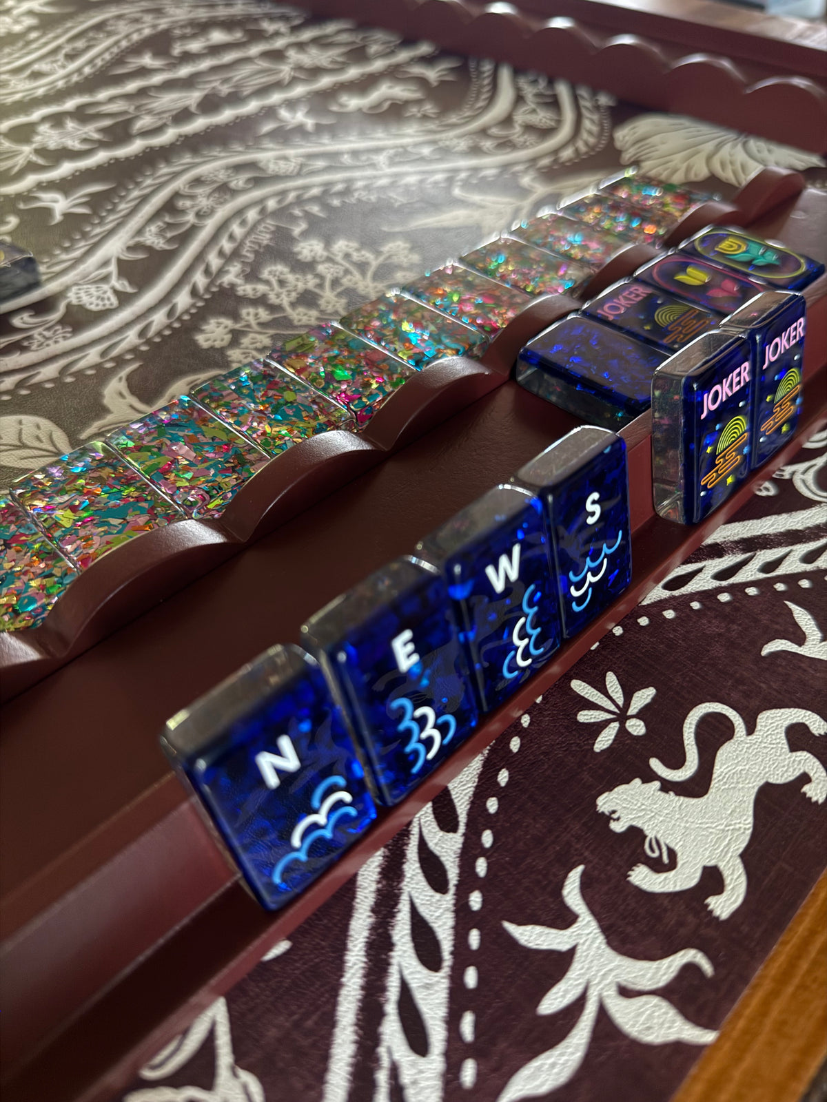 Close-up of a wind tile row and joker tiles resting in an oxblood mahjong rack with scalloped pusher, on tartan fabric