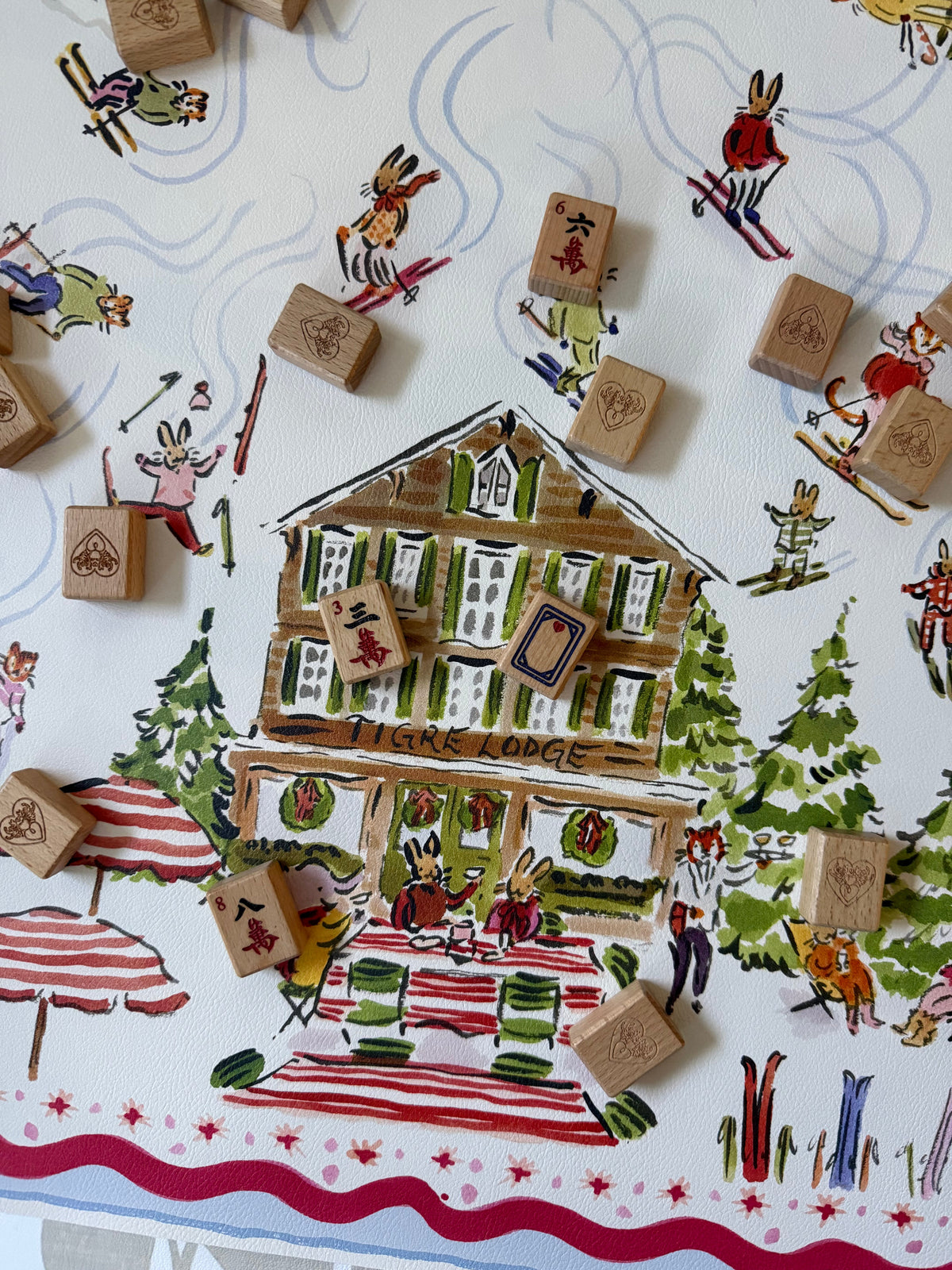 Paige Spearin Après Mahj mahjong mat: hand-illustrated Tigre Lodge ski chalet with skiing rabbits and tigers, wooden mahjong tiles scattered across the mat