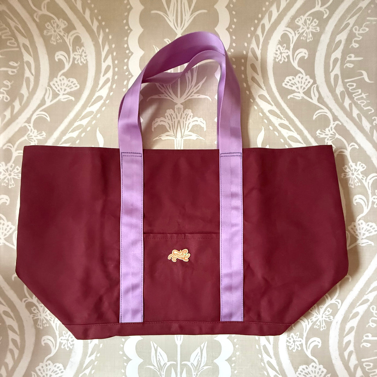 Signature XL Canvas Tote in oxblood canvas with lavender handles, front pocket and small embroidered tiger patch, laid flat