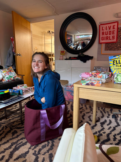 Woman smiling behind an oxblood Signature XL Canvas Tote with lavender handles in a living room