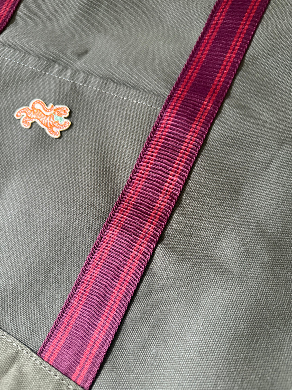 Close-up of the embroidered tiger patch and burgundy striped webbing handle on olive canvas