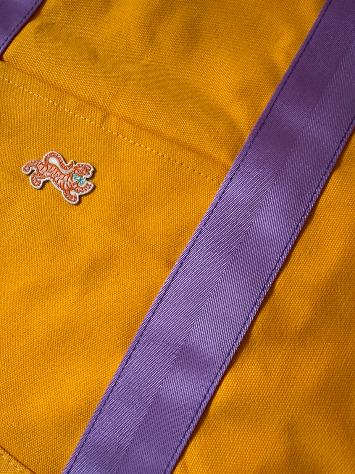 Close-up of the embroidered tiger patch and lavender webbing handle on marigold canvas