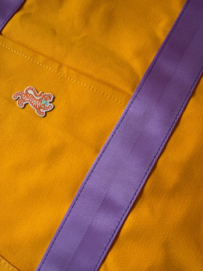 Close-up of the embroidered tiger patch and lavender webbing handle on marigold canvas