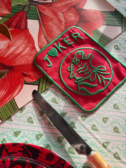 Corner of the Estate Sale mahjong mat with red amaryllis, under a red embroidered Joker coaster, a bamboo-handled knife and a red floral plate on a green-and-white tablecloth