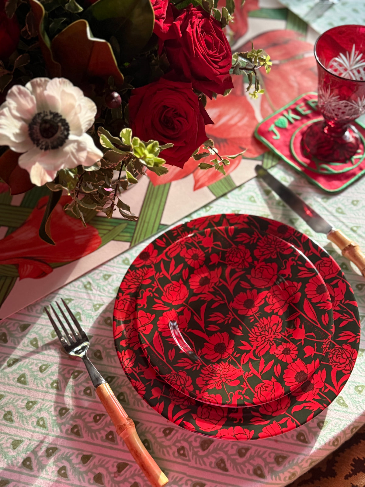 Estate Sale mahjong mat beneath a table setting of red floral plates, bamboo-handled cutlery, red roses and a white anemone
