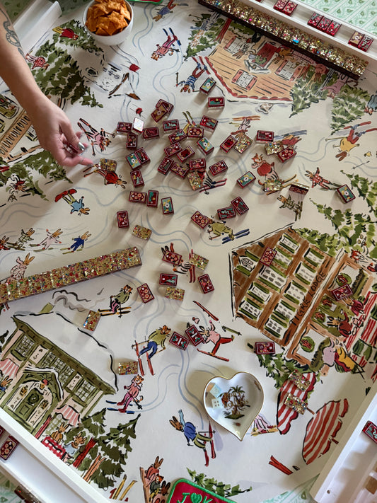 Paige Spearin Après Mahj mahjong mat mid-game, with tiles in the center, a hand reaching in, a heart-shaped dish and a bowl of snacks