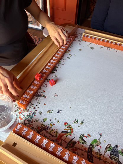Red logo dice rolled onto a white mahjong mat with an orange printed border, players racking tiles