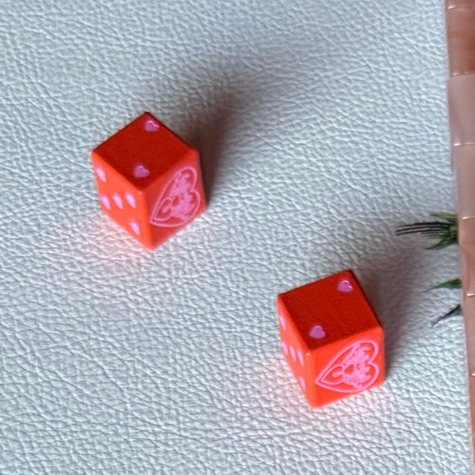 Two oversized red dice with pink heart pips and the Tigre de Tartán logo on a white faux-leather mat